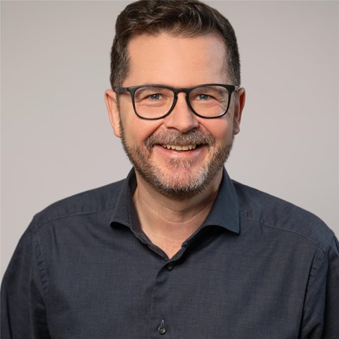 Ein Mann mit kurzen, dunkelbraunen Haaren trägt ein dunkelblaues Hemd mit offenem Kragen. Er steht vor einem einfarbigen, hellgrauen Hintergrund. Das Hemd hat eine Knopfleiste und ist schlicht ohne Muster oder sichtbare Accessoires.