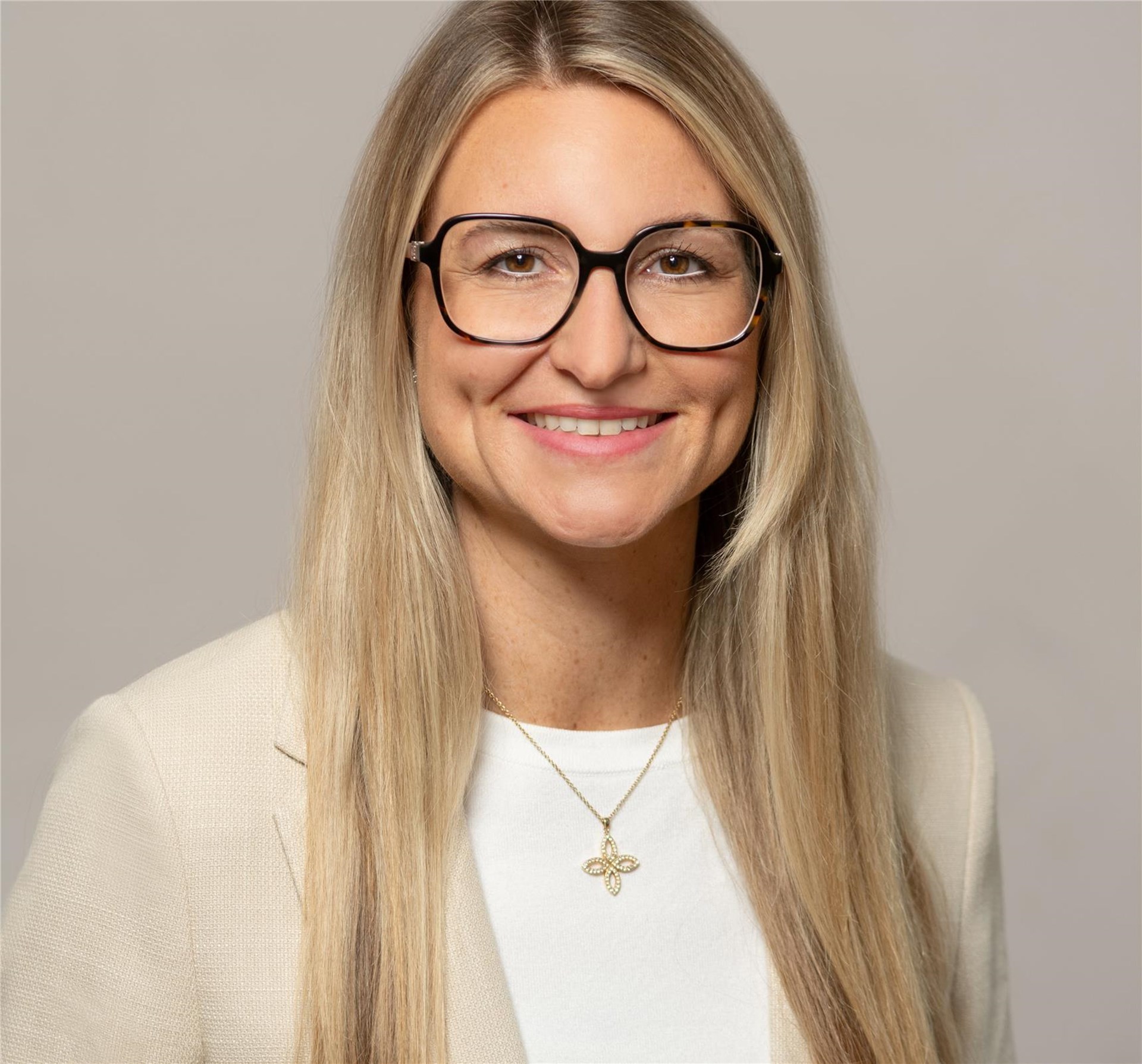 Frau mit langen blonden Haaren trägt hellbeige Blazer, weißes Shirt und goldene Kette mit kleeblattförmigem Anhänger.