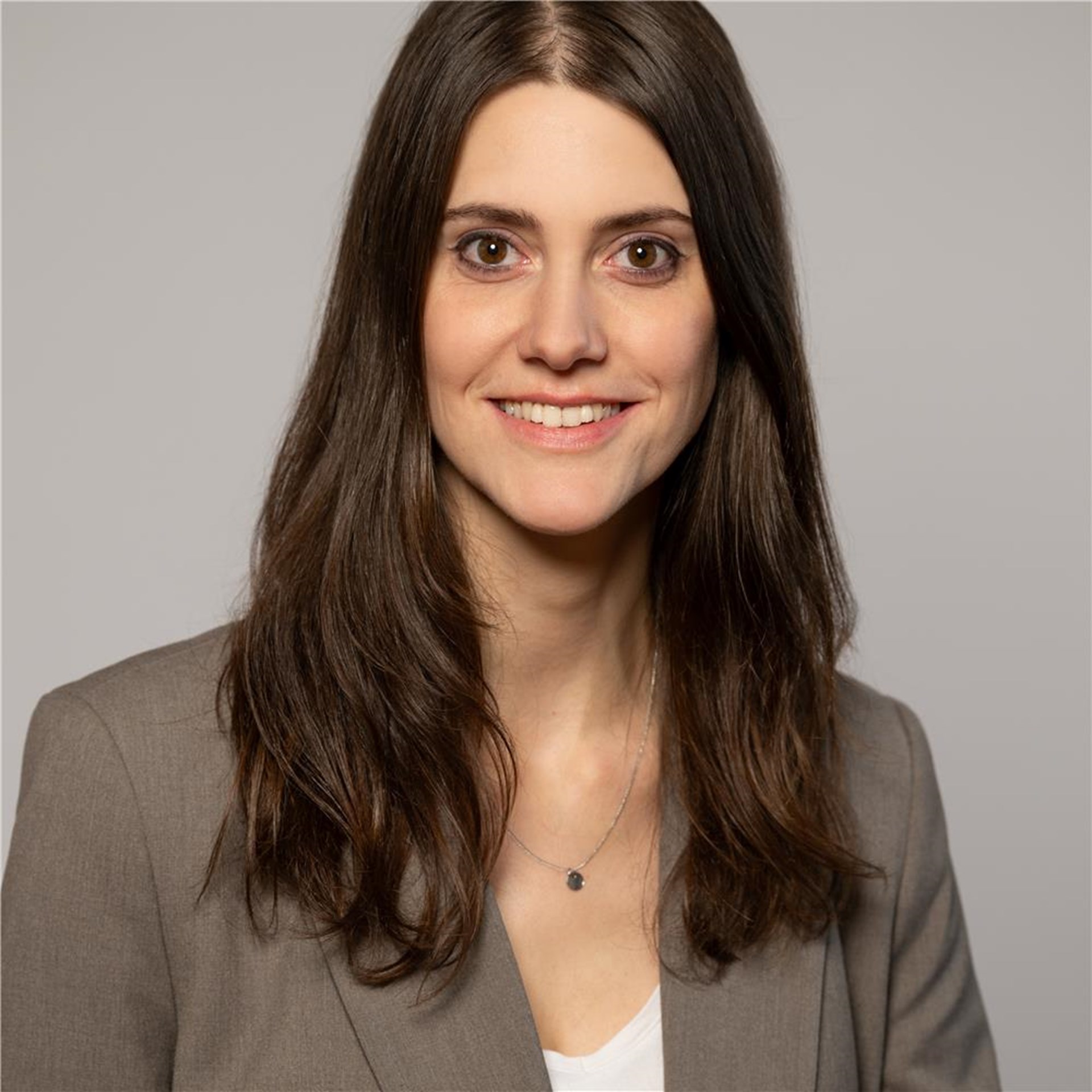 Frau mit langen braunen Haaren, trägt graue Blazerjacke, weiße Bluse und eine feine Halskette mit kleinem Anhänger.