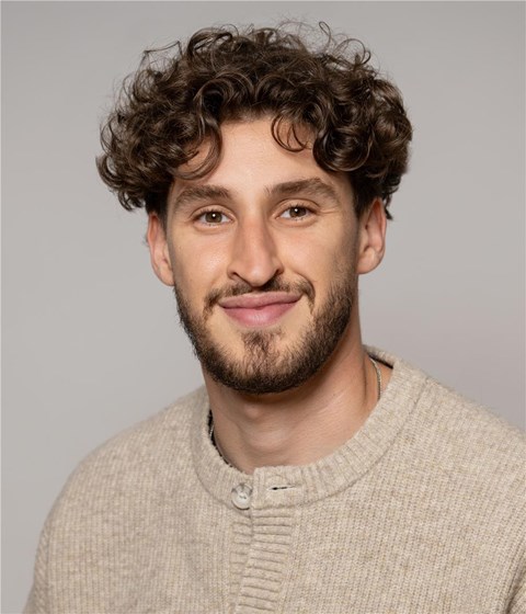 Das Bild zeigt einen lächelnden Mann mit braunen lockigen Haaren und beigem Pullover.