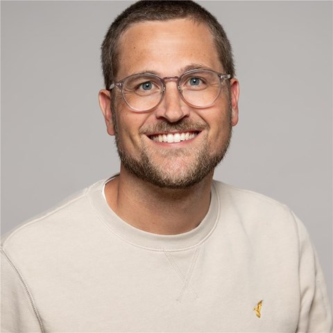 Ein Mann mit kurzen braunen Haaren, dreitagebart, Brille und lächelt in die Kamera, er trägt einen weißen Pullover